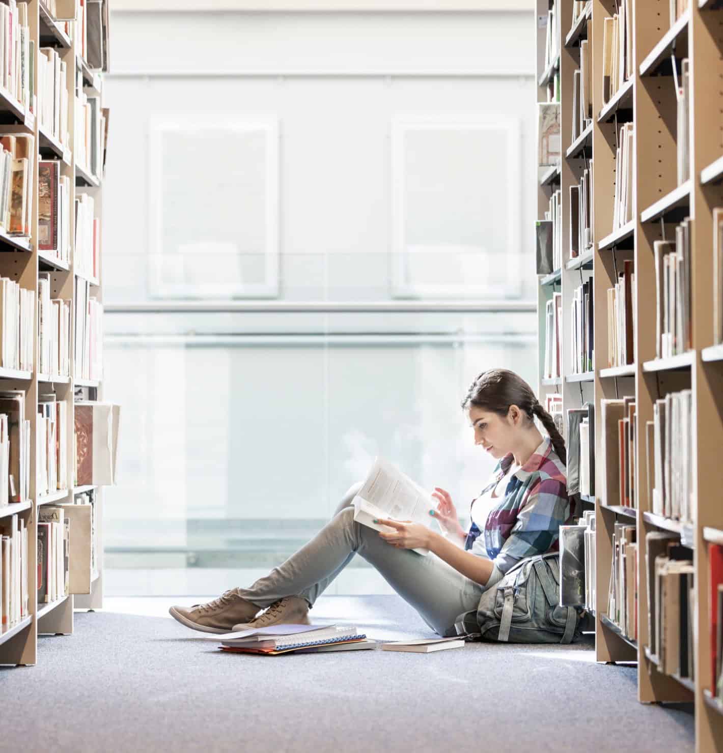 A girl reading a book sitting on the floor inbetween bookshelves at the library.